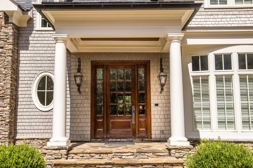 Front entrance of a house with tall white columns, gray shingle siding, and elegant custom iron doors. The wooden double door with glass panes, two lantern-style lights, stone steps, and large white-framed windows create timeless charm in Middlesex & Bergen County, NJ.