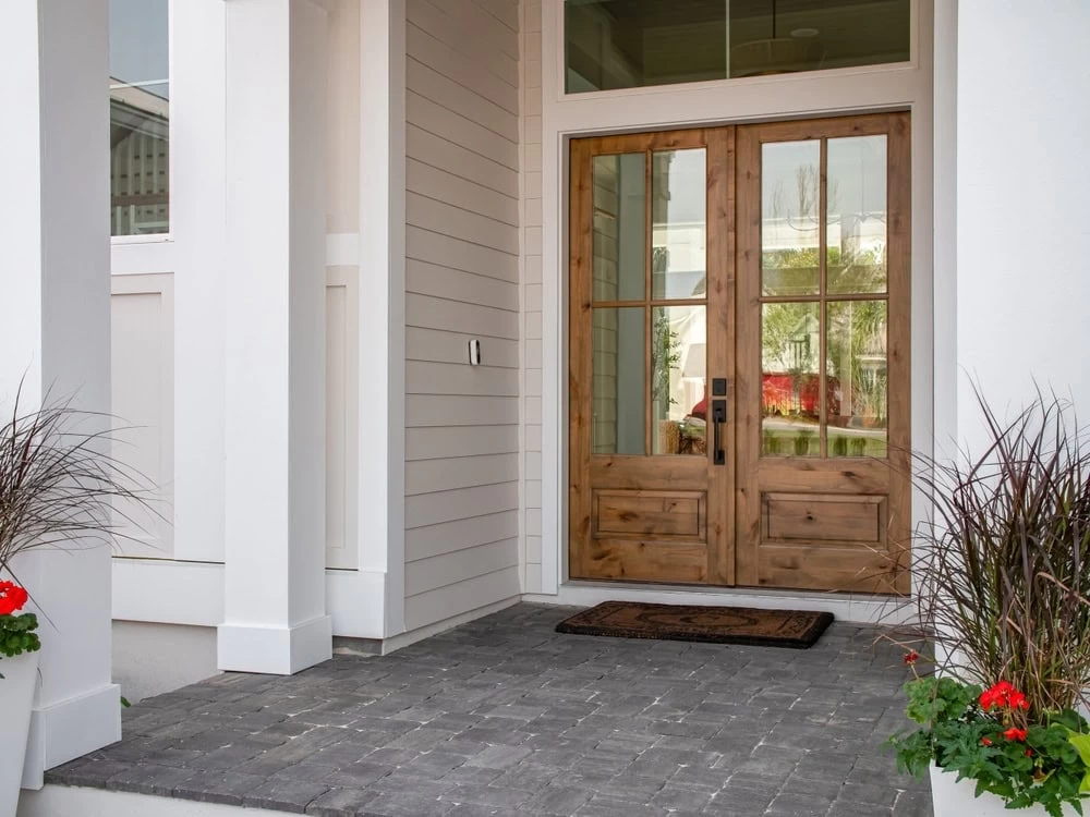 A modern front porch with custom iron doors in Middlesex & Bergen County, featuring glass panels, white siding, gray stone steps, a patterned welcome mat, and planters with red flowers and tall grasses on either side.