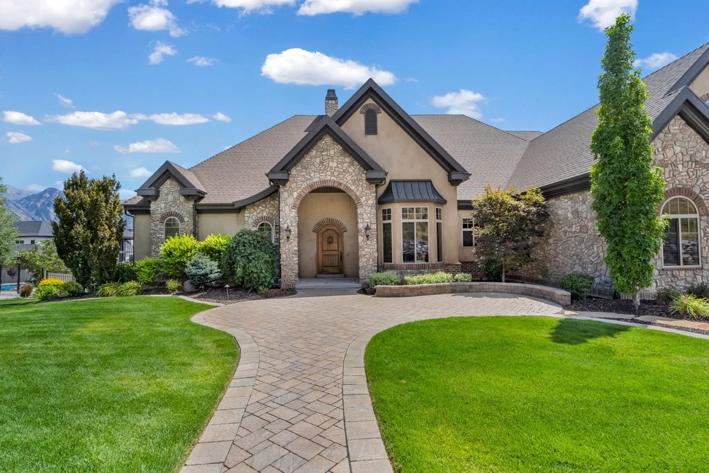 A stone and stucco house with arched entryway featuring custom iron doors Middlesex & Bergen County, NJ, manicured lawn, curved brick pathway, and landscaped bushes under a blue sky with scattered clouds.