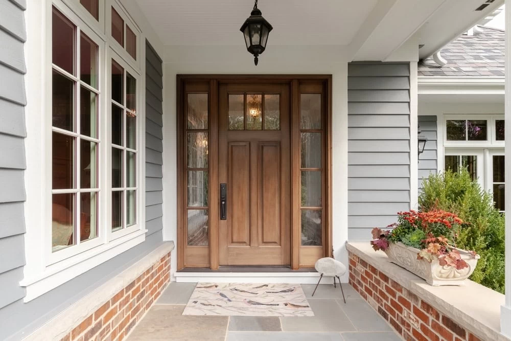 A welcoming front porch with large windows, gray siding, and red brick accents. A pastel doormat, potted plants, and a small stool decorate the entrance. For NJ elegance, consider custom iron doors Middlesex & Bergen County above the black lantern.