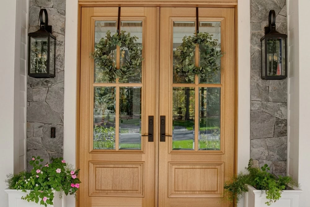 Double wooden front doors with glass panels, each adorned with a green wreath. Stone wall background, black outdoor lanterns, and white planters frame the entrance—perfect inspiration for custom iron doors in Middlesex & Bergen County, NJ.