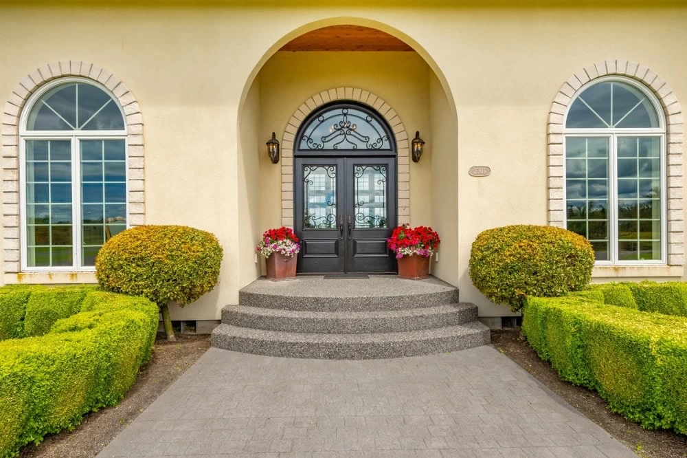 Elegant front entrance featuring custom iron doors Middlesex & Bergen County, NJ, with ornate glass, arched windows, potted flowers, trimmed bushes, and manicured green hedges lining the curved pathway.