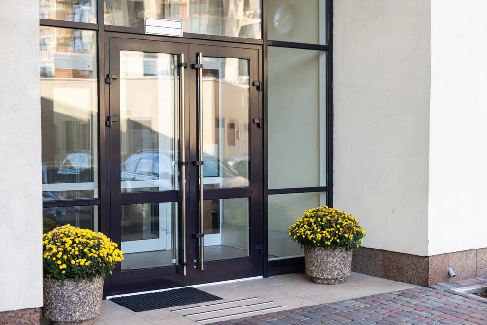 A glass double door entrance, flanked by two large pots of yellow flowers, welcomes visitors. Reflections of parked cars shimmer in the glass, and a doormat awaits beneath. Explore custom iron doors in Middlesex & Bergen County, NJ for a touch of elegance.