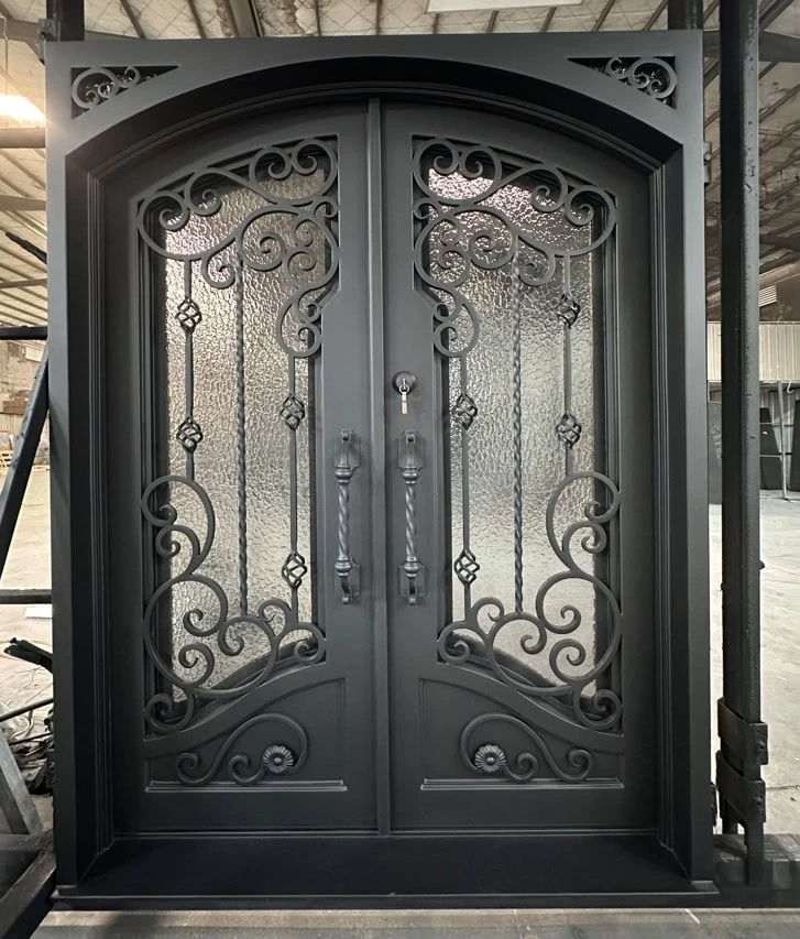 A pair of ornate black double doors with decorative wrought iron scrollwork and textured glass panels, set in an arched black frame inside a warehouse space.