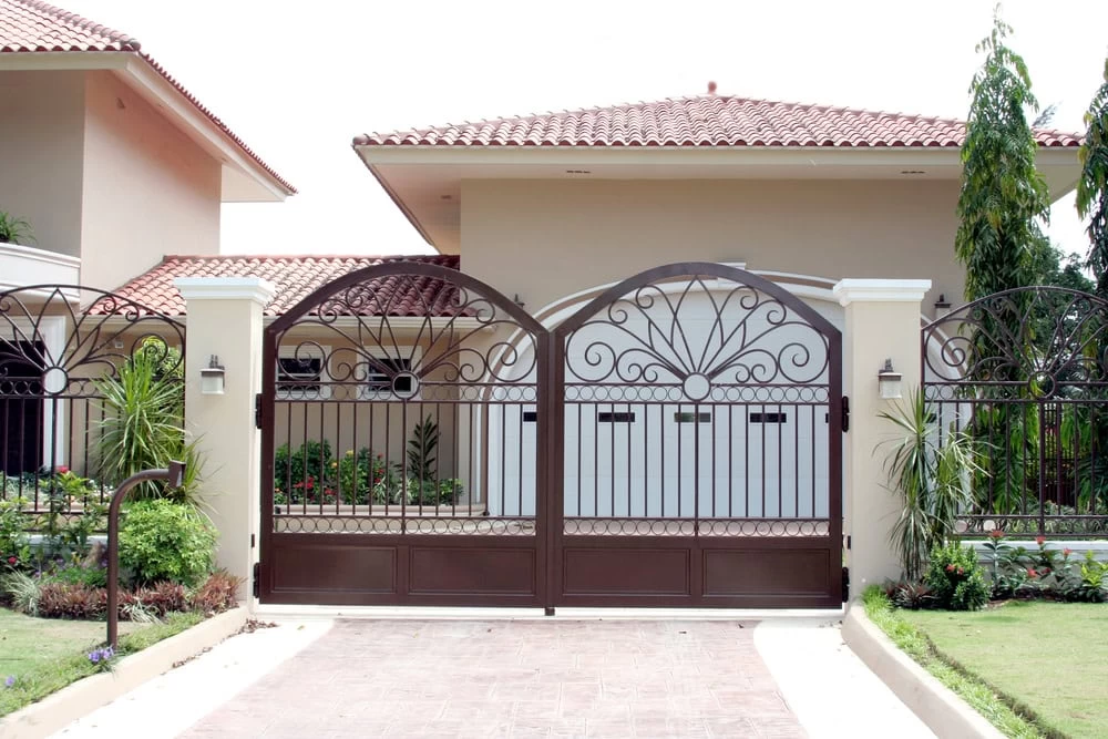 A modern house with a red-tiled roof stands behind tall decorative iron doors in Middlesex & Bergen County, NJ, framed by landscaped greenery, stone pillars, and a paved driveway leading to the gate.