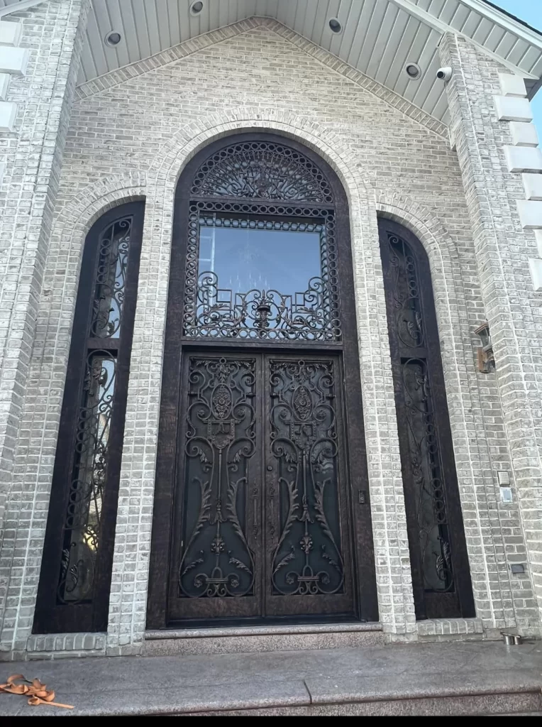 Ornate double front doors with intricate wrought iron designs, flanked by tall matching side panels and topped with a large arched window, set in a light-colored brick facade.
