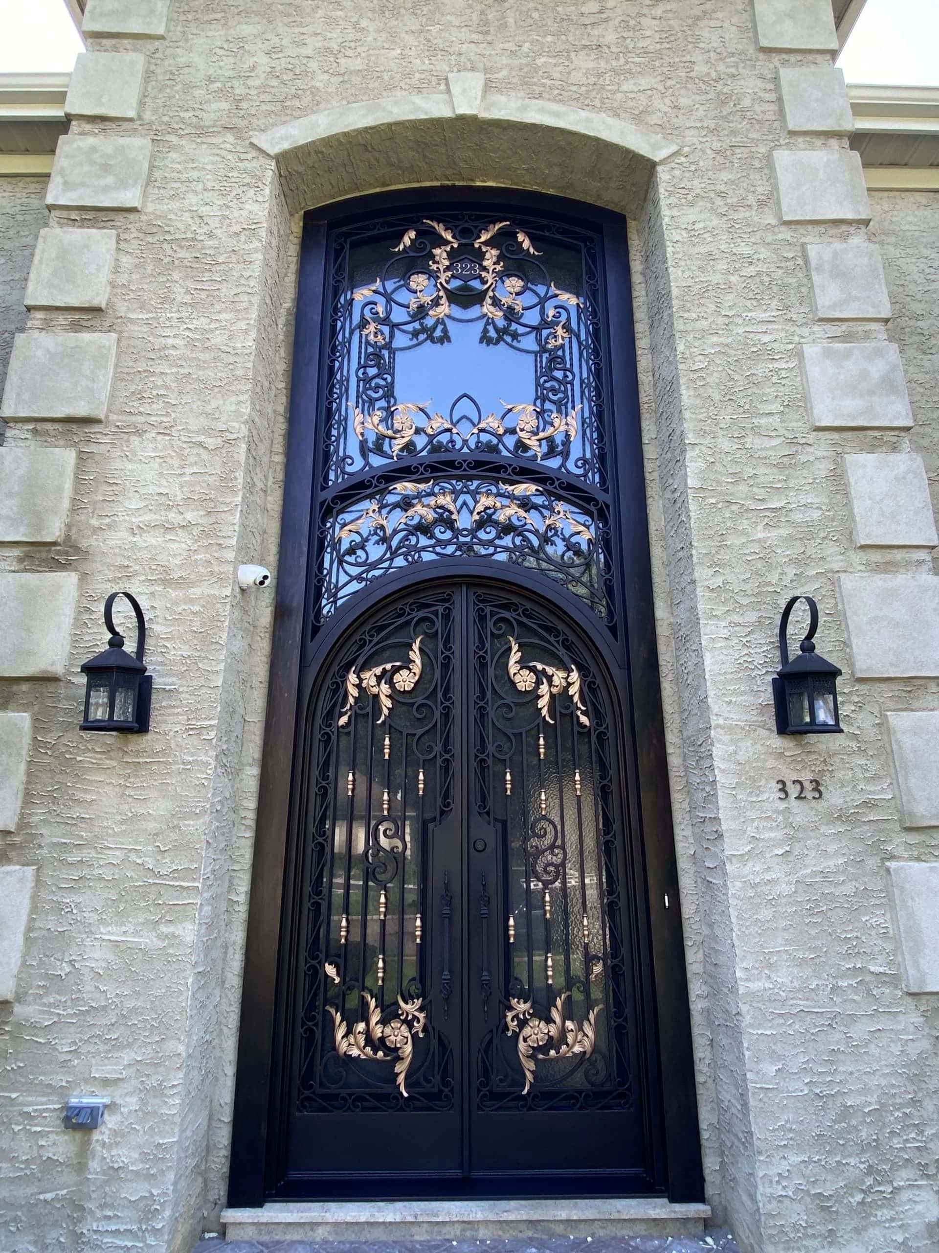 Tall, ornate black double doors with gold decorative accents and intricate ironwork designs, set in a textured beige stone exterior. Two black lantern-style lights flank the entrance, and the number 323 is displayed on the wall.