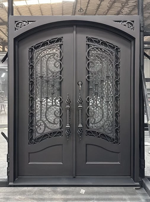 Ornate double iron doors with decorative scrollwork and glass panels stand upright in a warehouse setting, featuring intricate patterns and two matching handles.