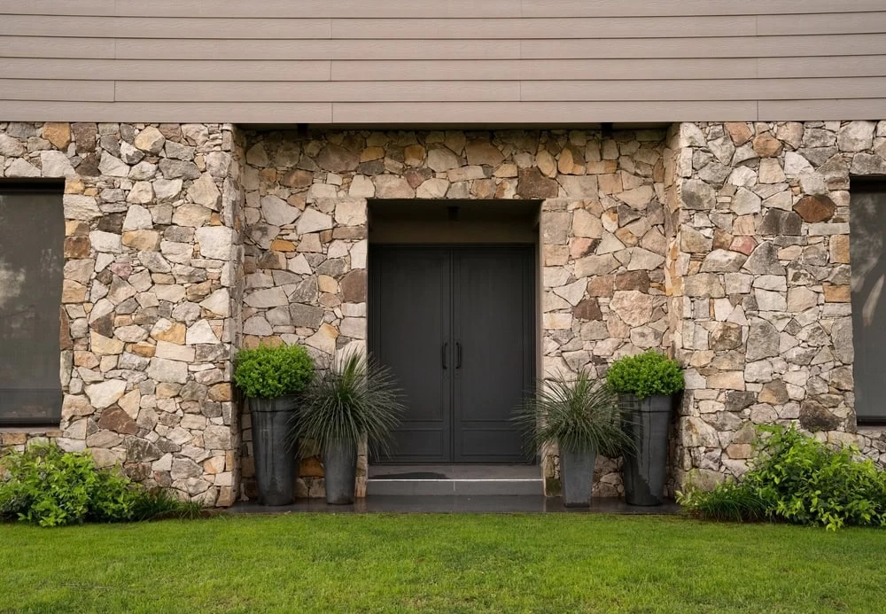 A house entrance with custom iron doors in Middlesex & Bergen County, NJ, set in a stone wall, flanked by tall gray planters holding green shrubs and grass, with manicured lawn and bushes in front.
