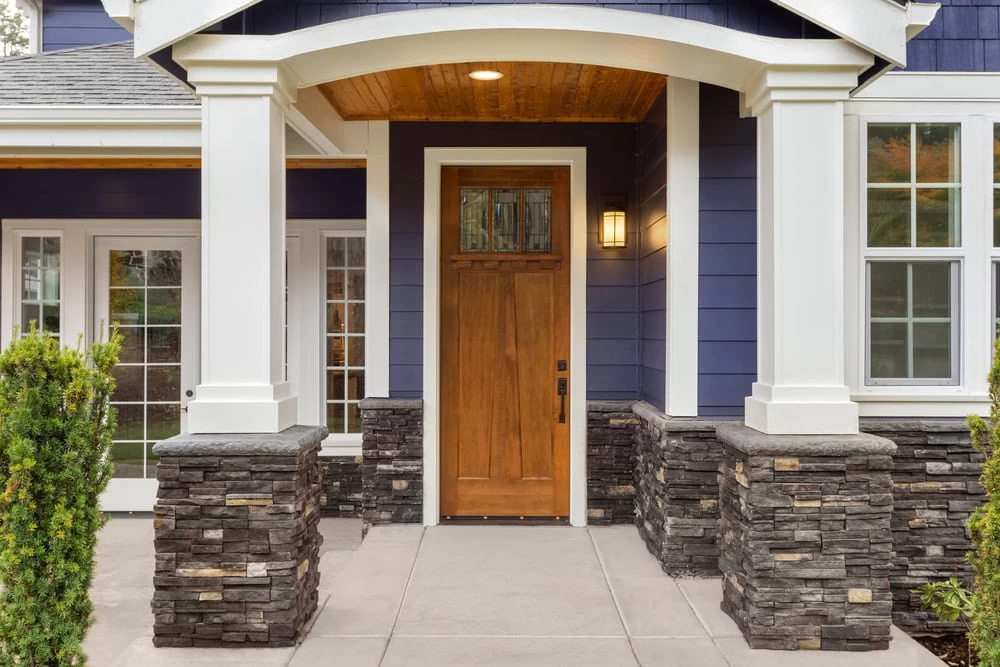 Front entrance of a house with a wooden door, stone columns, white trim, blue siding, and a covered porch ceiling with wood accents—showcasing the classic style found in homes with iron doors Middlesex & Bergen County residents admire.