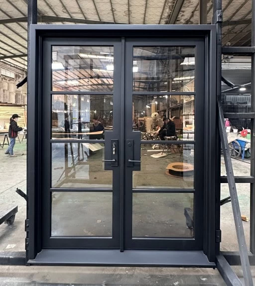 A pair of modern black-framed glass double doors are installed in an industrial workshop, with people and equipment visible in the background through the glass.