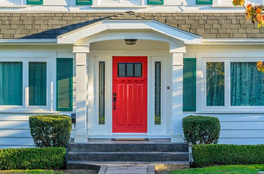 Front view of a white house in NJ with teal shutters and a striking red door, framed by trimmed bushes and a small porch—perfect for showcasing custom iron doors Middlesex & Bergen County residents love.