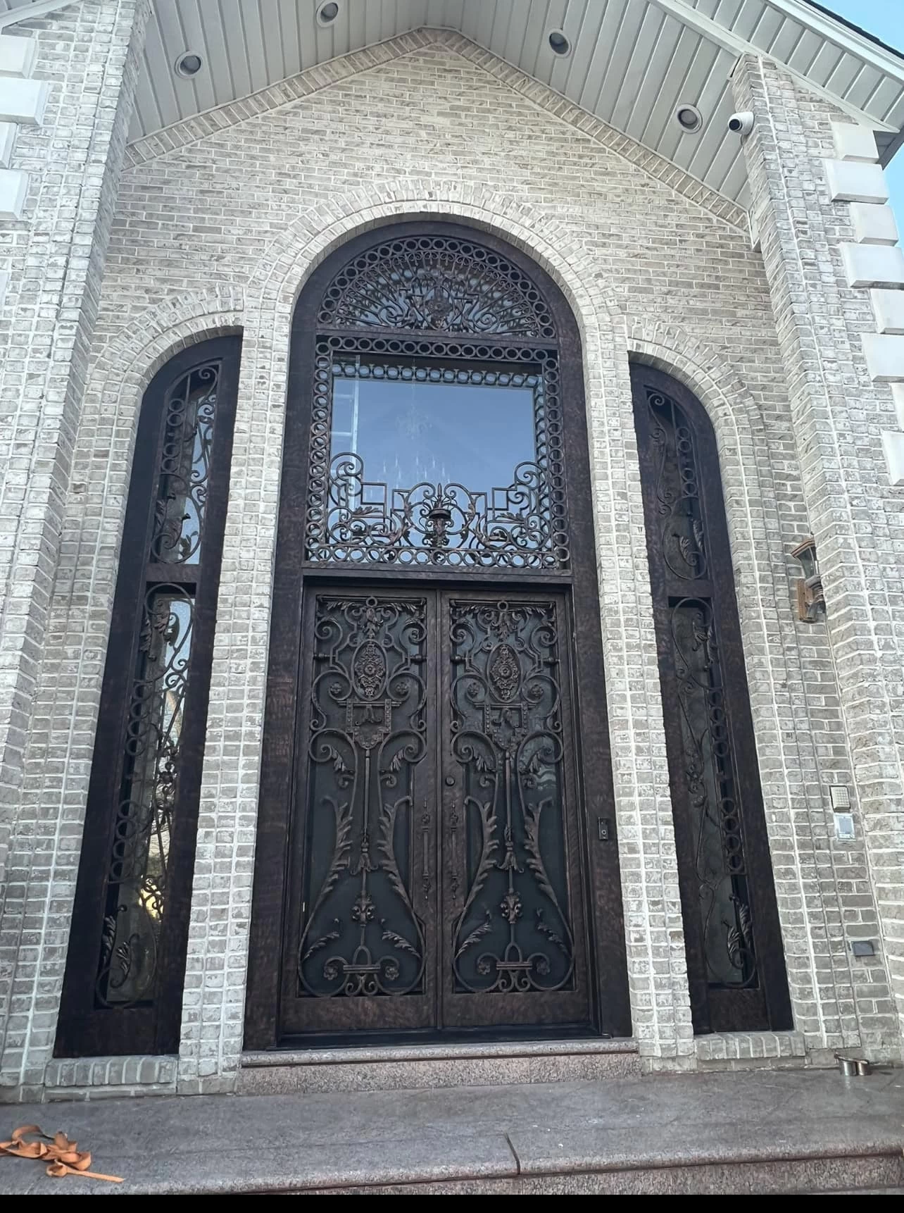 Ornate double front doors with intricate wrought iron designs, flanked by tall matching side panels and topped with a large arched window, set in a light-colored brick facade.