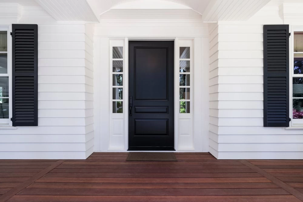 A modern, black front door with sidelights is centered on a white house with black shutters and wood plank flooring on the porch.