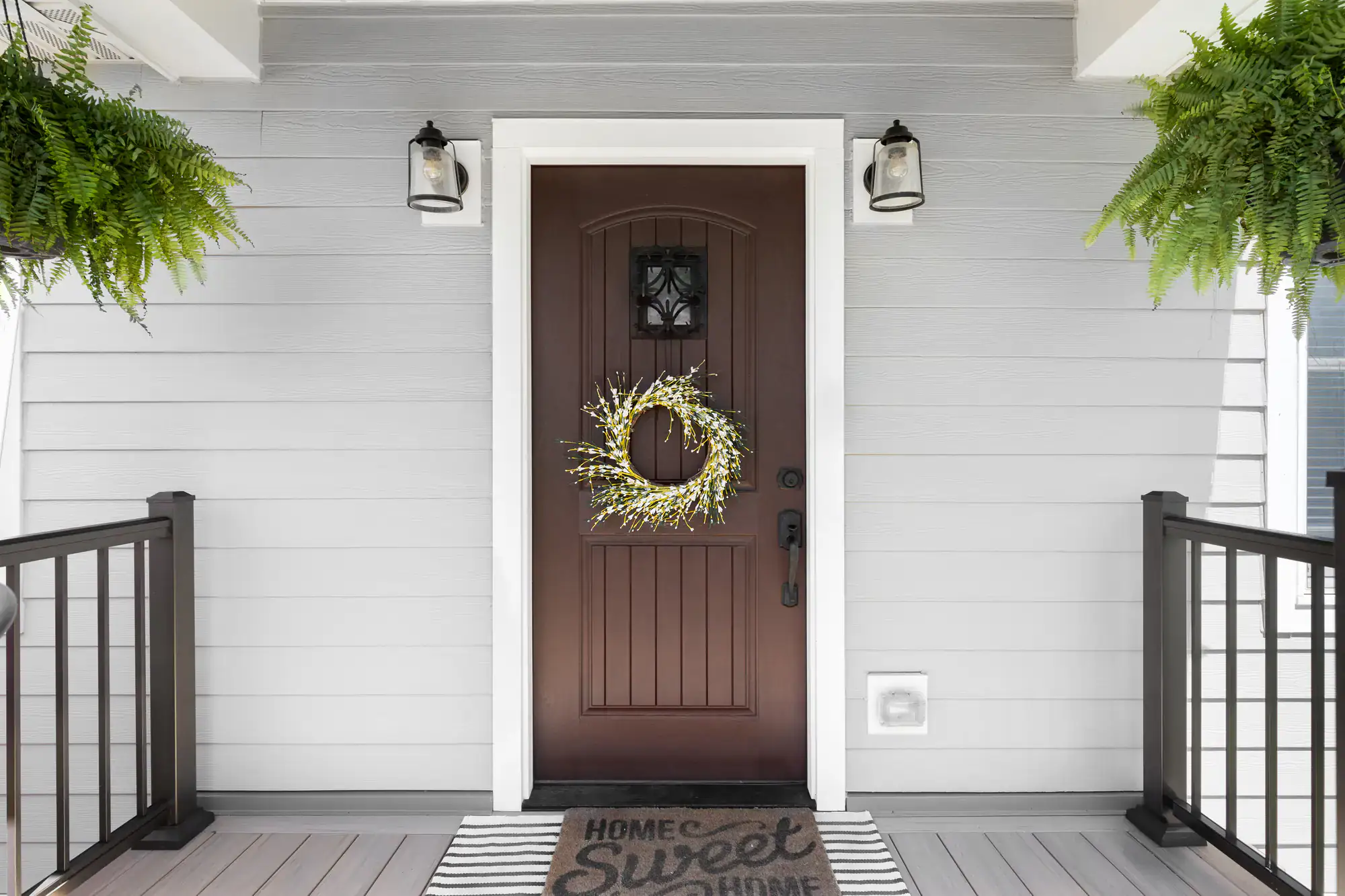 A front porch with a dark wooden door decorated with a yellow floral wreath. The door, ideal for homes in Bergen County, is flanked by lantern-style lights, hanging ferns, and a “Home Sweet Home” doormat on a striped rug. White siding and black railings complete the scene.