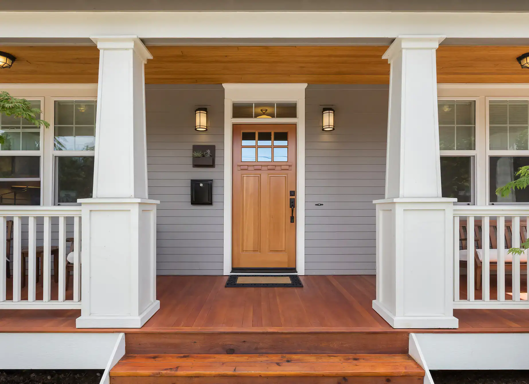 A front porch with wooden steps, white railing and columns, a light gray exterior, and an elegant iron door with glass panes—perfect for homes in Bergen County—flanked by two windows and two wall-mounted lantern lights.