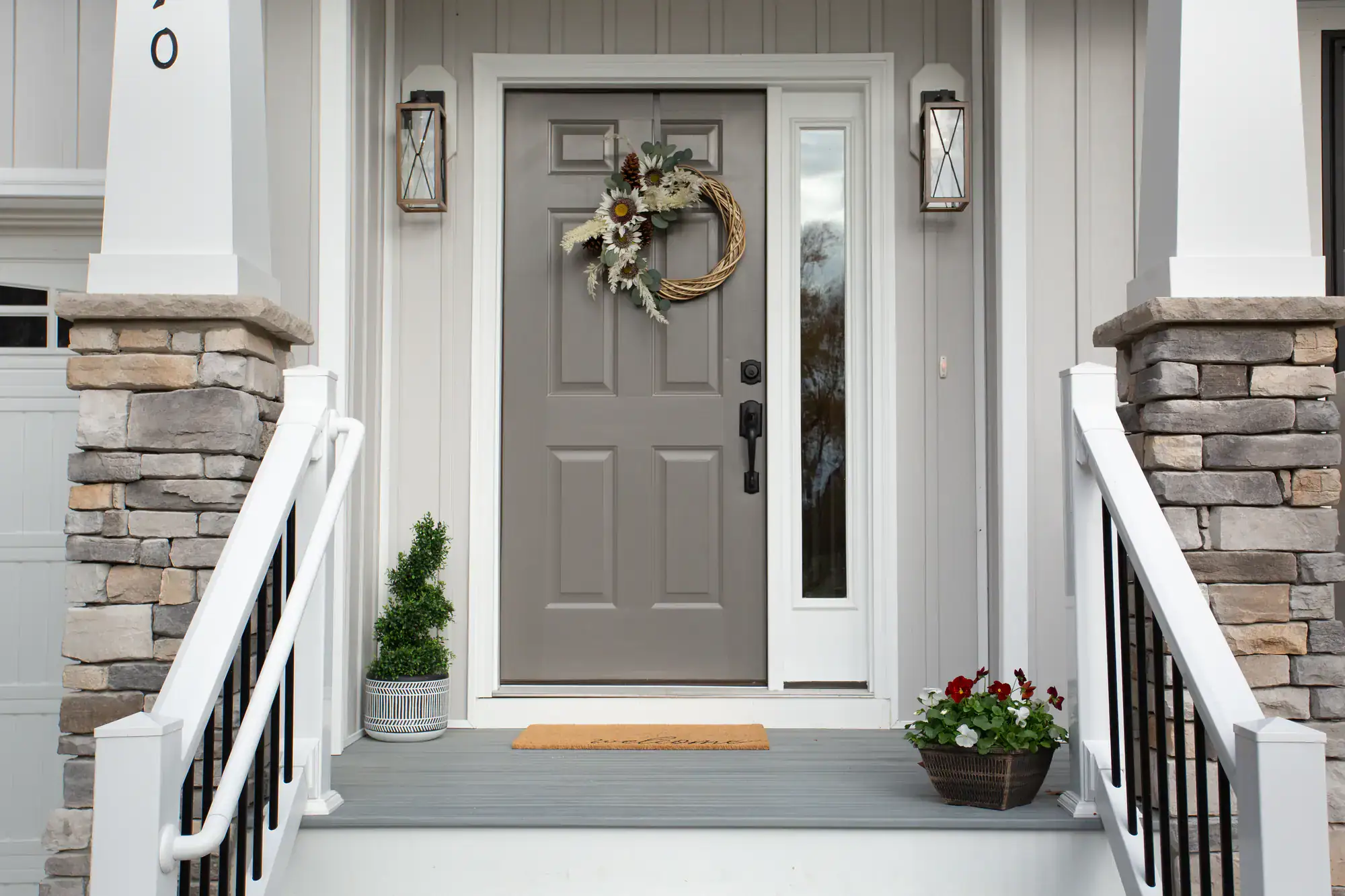 A gray front door with a decorative wreath, flanked by two lantern-style lights, welcomes you in Bergen County, NJ. Stone columns, white railings, a doormat, and a flower pot add charm to this inviting porch—explore more than just iron doors Middlesex.