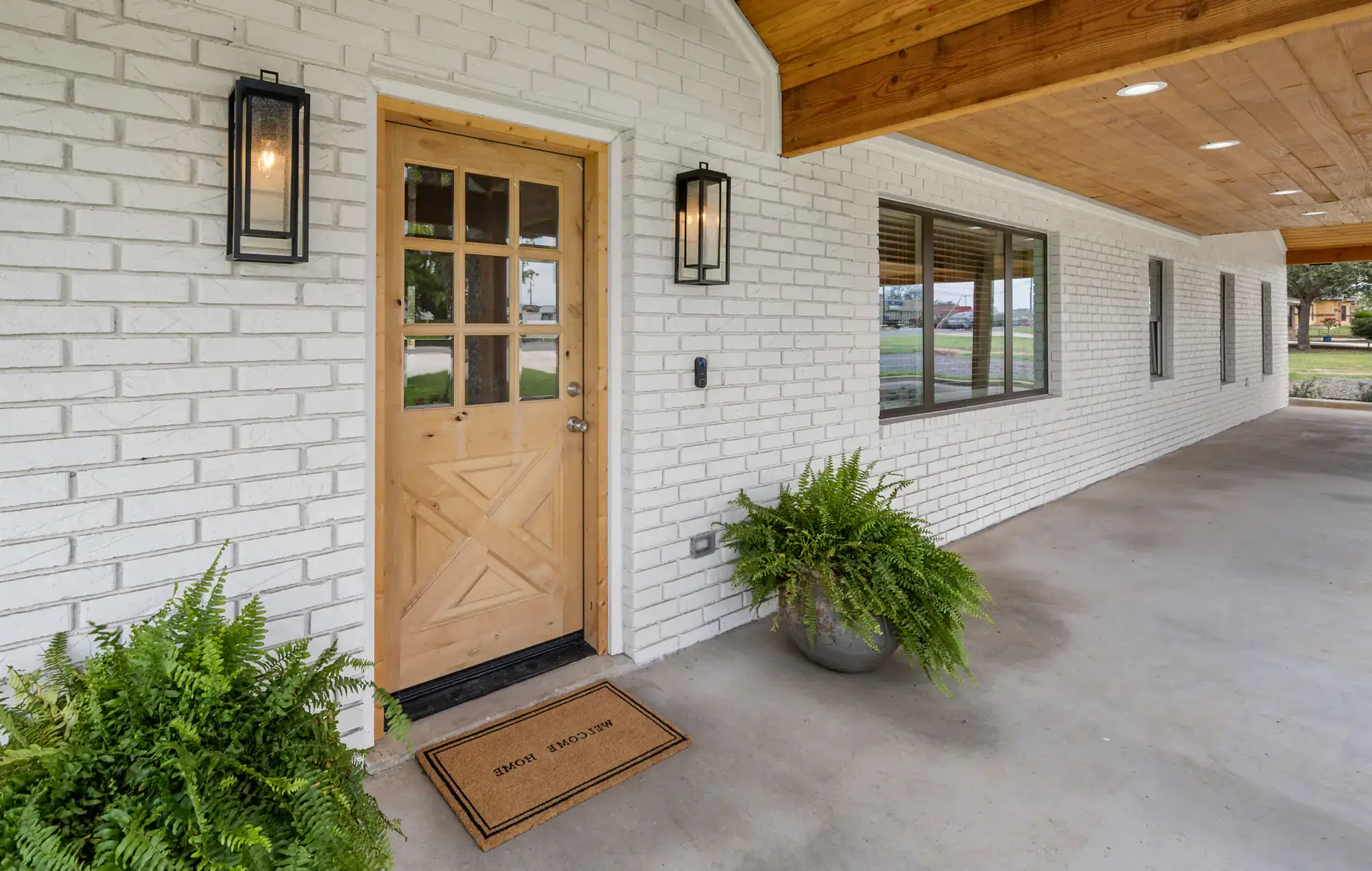 A light wooden front door with a glass panel is set in a white brick wall, creating classic Bergen County charm. Two black lantern lights and green potted ferns flank the entrance, while a “Welcome Home” doormat lies on the concrete porch.